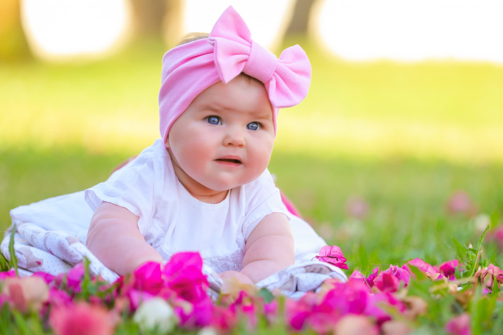Baby girl with a pink bow headband lying among bright petals
