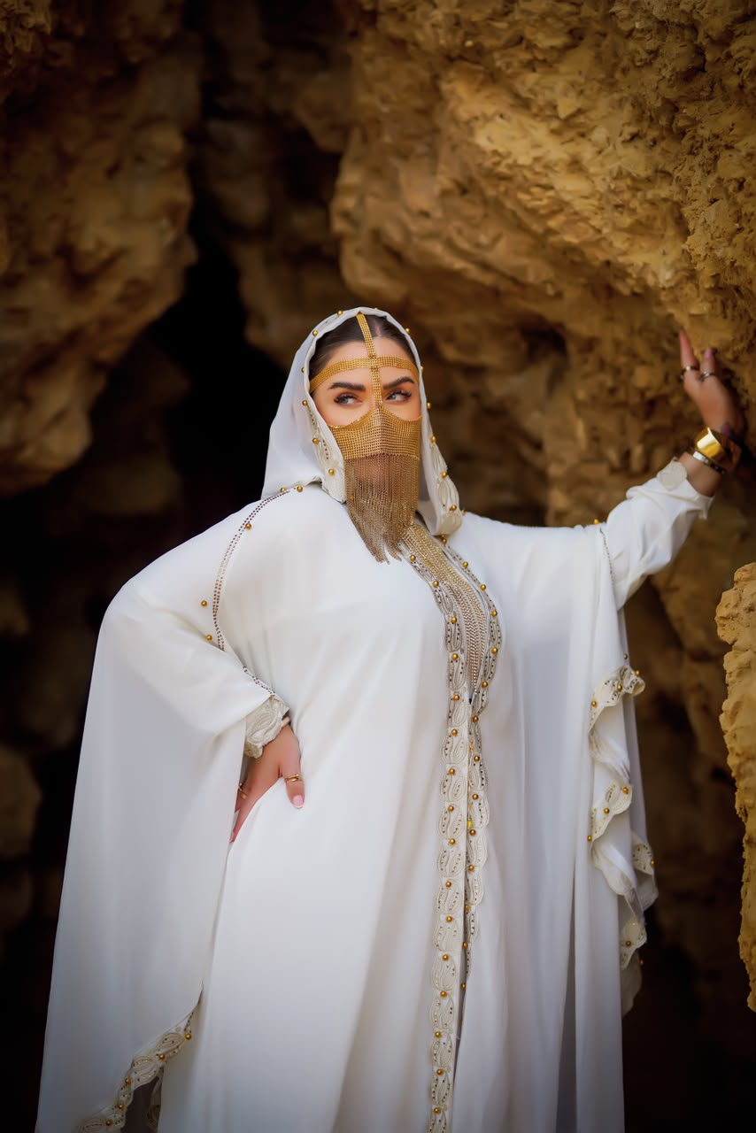 Woman in a white abaya and gold veil posing inside a sandstone cave