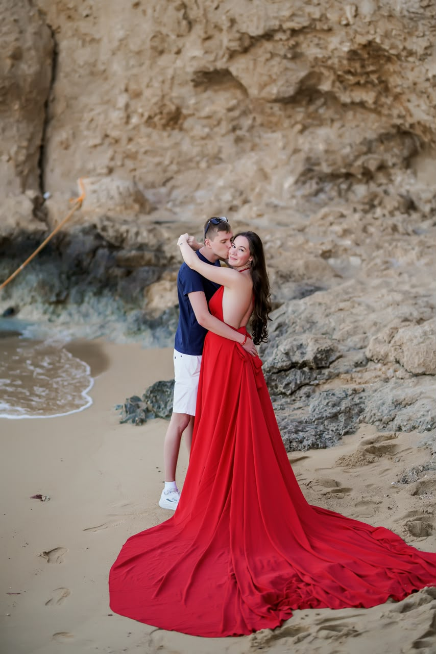 Couple kissing on the sand, red gown train spread across the beach