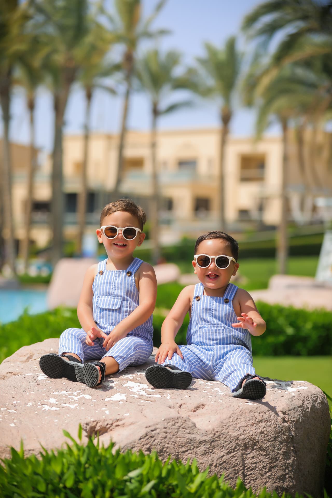 Two toddlers in striped dungarees and sunglasses sitting on a rock