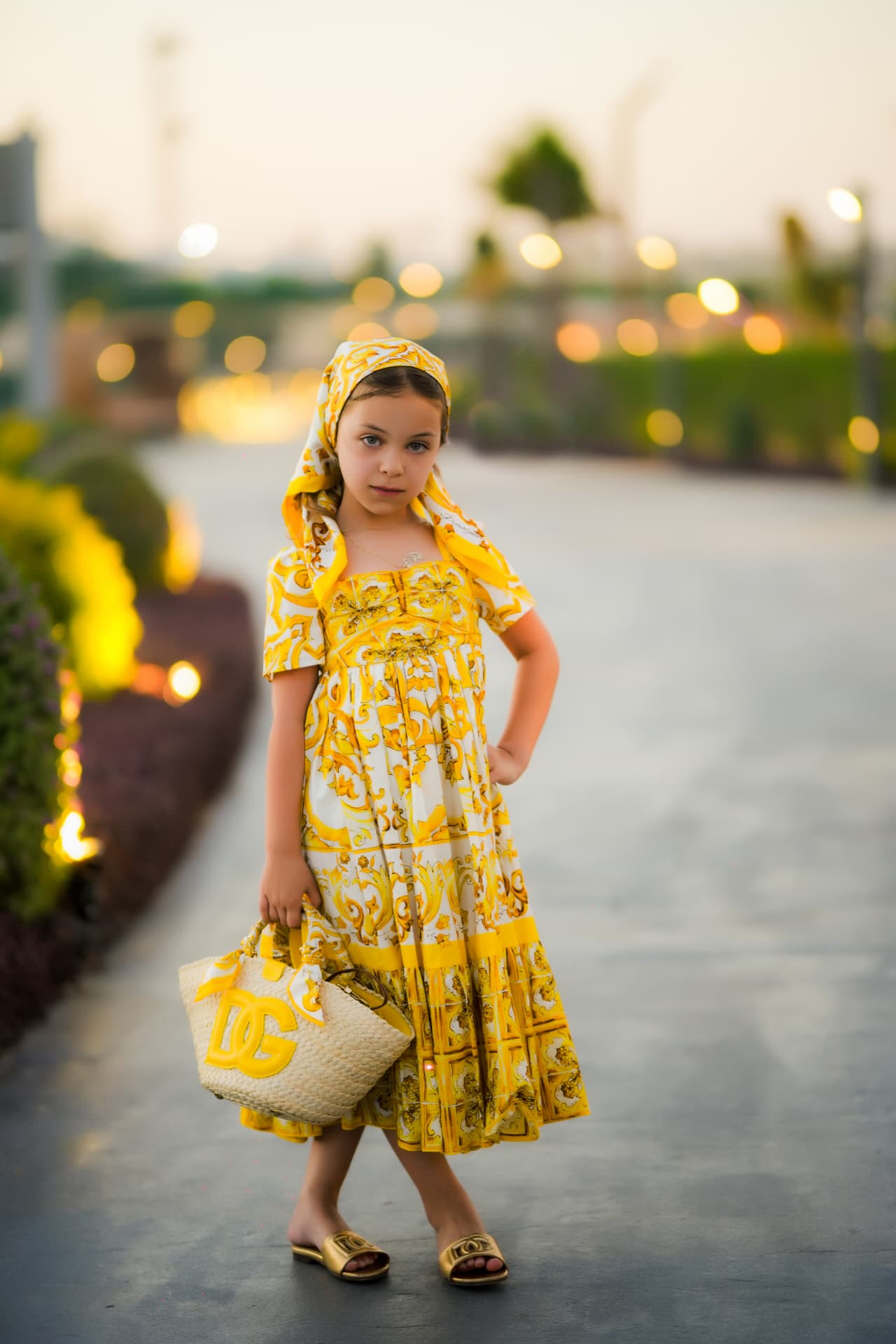 Girl in a yellow print dress and matching headscarf on a lit walkway