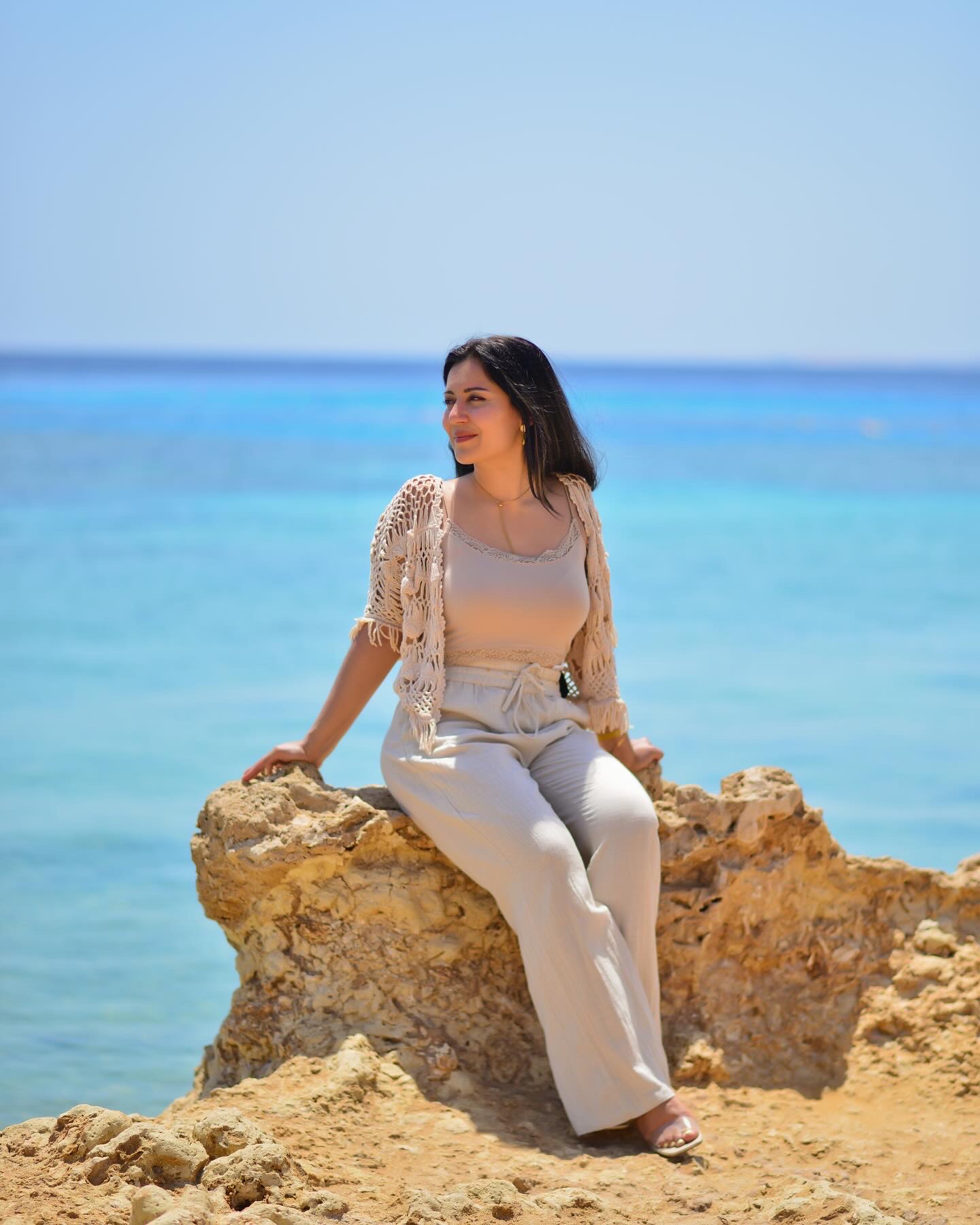 Woman in beige linen seated on a rock above the blue sea