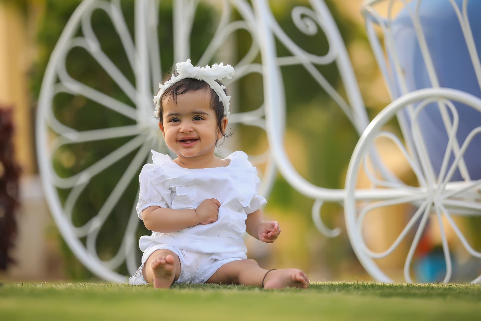 Baby girl in white sitting on the grass beside a white carriage prop