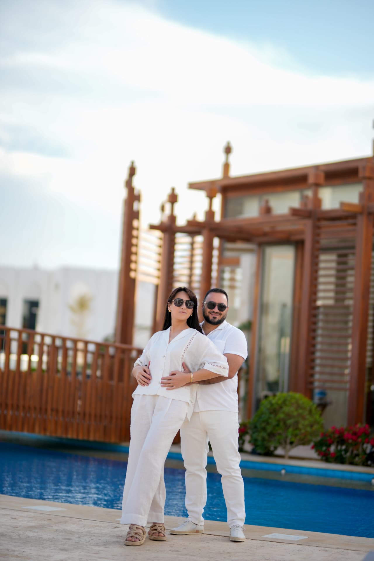 Couple in white outfits posing by a poolside pergola